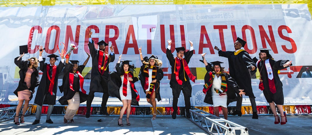 students in regalia jumping in front of congratulations sign